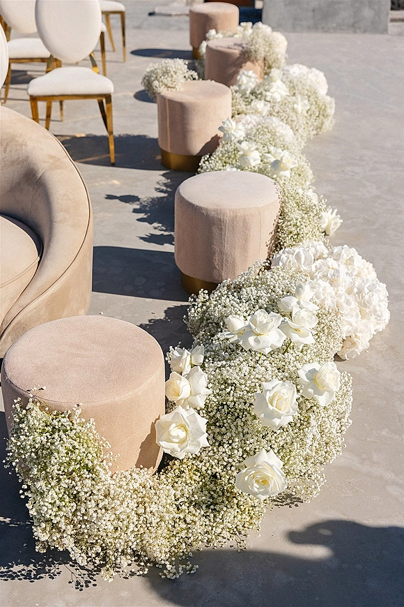 Ceremony aisle decor with wedding aisle flowers of white roses, baby’s breath, and hydrangeas beside blush velvet ottomans on a patio
