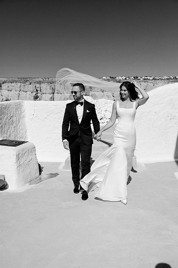 Couple portrait, black and white wedding portrait of bride and groom holding hands by white stone walls on a cliffside under clear sky