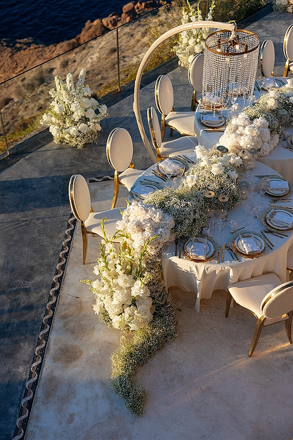 Reception tablescape with round wedding table setup, white florals and gold flatware beneath a chandelier arch on a cliffside patio by ocean
