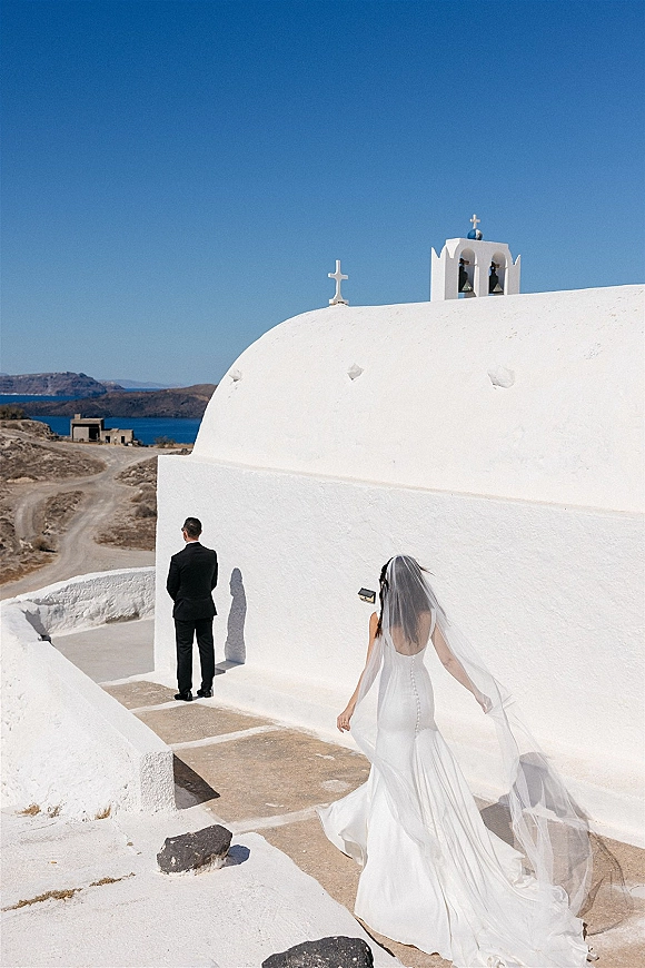 Wedding first look as the bride in a strapless gown and long veil walks up to the groom in a black suit outside a white seaside chapel