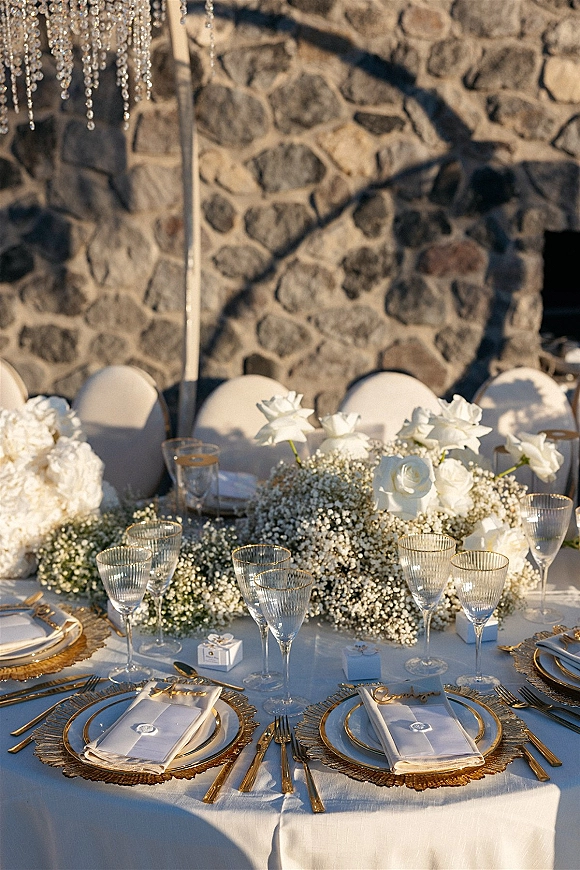 Reception tablescape with white and gold tablescape details, white rose centerpiece, gold flatware, ribbed glasses, beneath crystal chandelier by stone wall outdoors