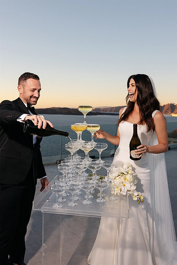 Champagne tower toast as bride and groom pour into coupe glasses on an acrylic table, veil and tuxedo framed by ocean cliffs backdrop
