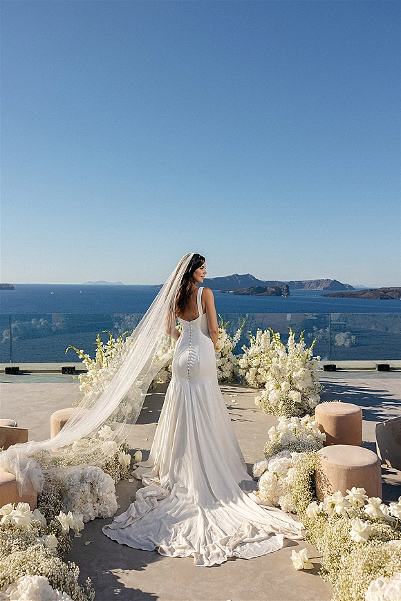 Bridal portrait of bride back view in a simple satin wedding dress with long cathedral veil on a terrace overlooking ocean and islands