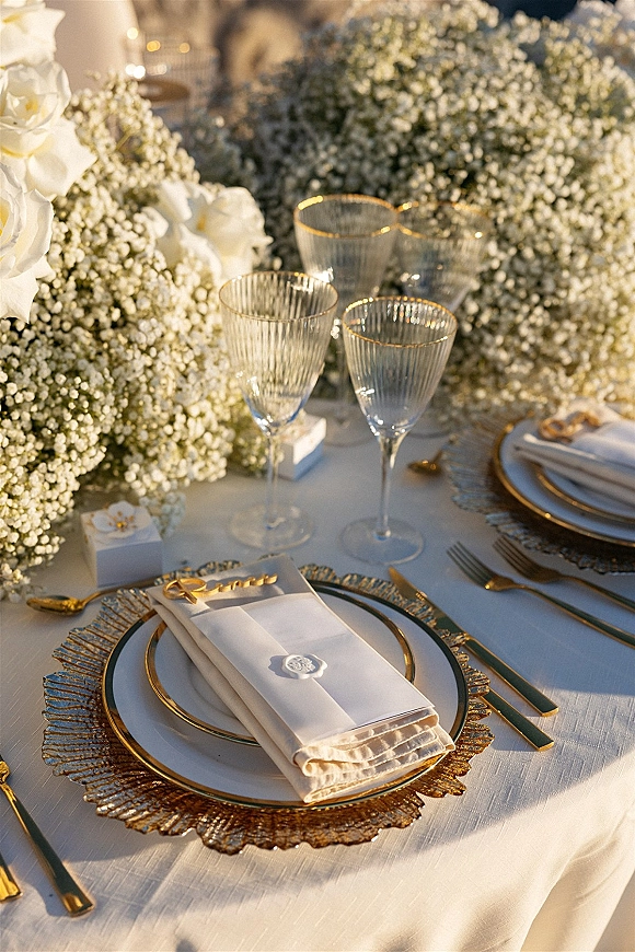Reception tablescape with wedding place setting featuring white roses and baby's breath, gold charger plates, and wax seal name cards in warm sunlight