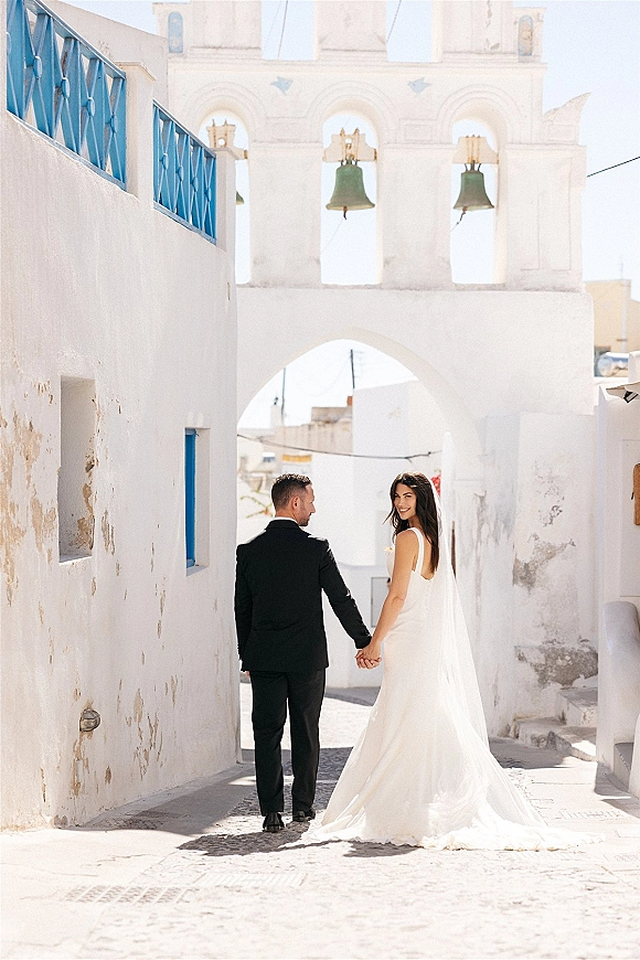 Couple portrait of bride and groom holding hands walking down a cobblestone street, bride looking back with veil, near white stucco bell tower