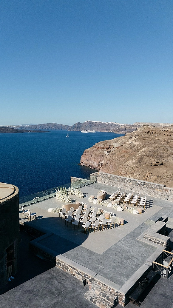 Ceremony setup for an outdoor wedding ceremony with round chairs and white floral pedestal stands on a stone terrace overlooking ocean cliffs