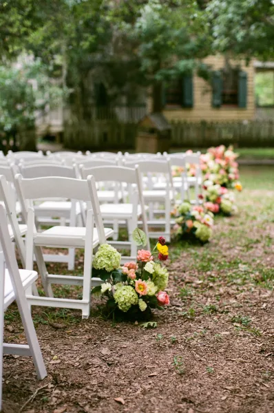 Ceremony aisle design with outdoor ceremony seating lined by hydrangeas along a dirt path on a garden lawn bordered by garden trees