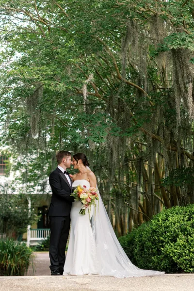 Couple portrait of a bride and groom embrace, foreheads touching, as she holds a colorful bouquet beneath garden trees draped with Spanish moss.