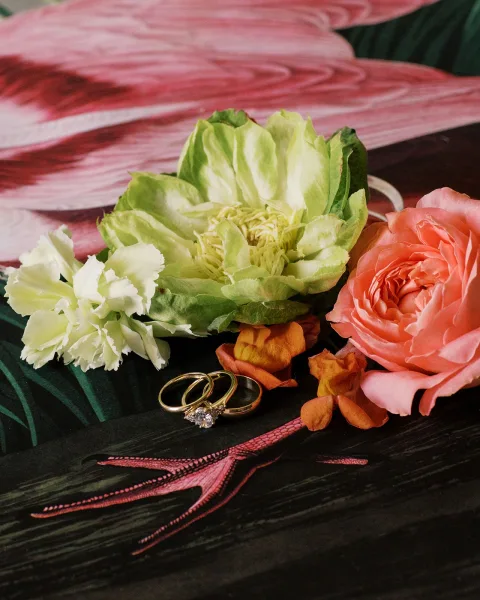 Wedding rings with a diamond engagement ring styled among roses on a dark, patterned surface, arranged as a refined flat lay.