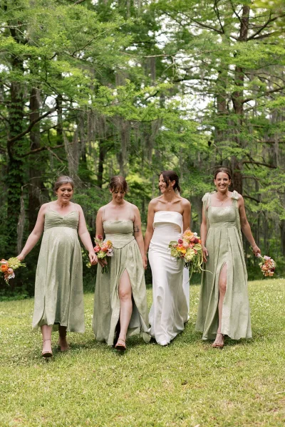 Bride and bridesmaids share a joyful bridal party portrait, bouquets in hand on a lawn beneath moss-draped trees in soft light.