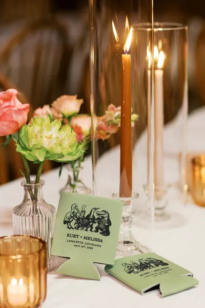 Wedding koozies and personalized can coolers arranged on a white tablecloth, framed by taper candles with wooden chairs in the background.