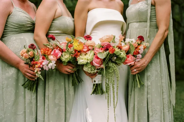 Bridesmaid bouquets frame this bridal party bouquets lineup, ribbon-tied in peach, pink, coral, and yellow, against garden trees.