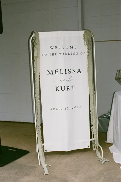 Wedding welcome sign on a fabric wedding banner hung from a metal stand, presented simply to greet arriving guests at the celebration.
