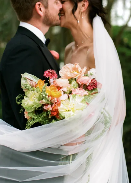 Wedding kiss portrait of a couple wrapped in veil, faces close amid garden trees and greenery, capturing a tender, romantic moment together.