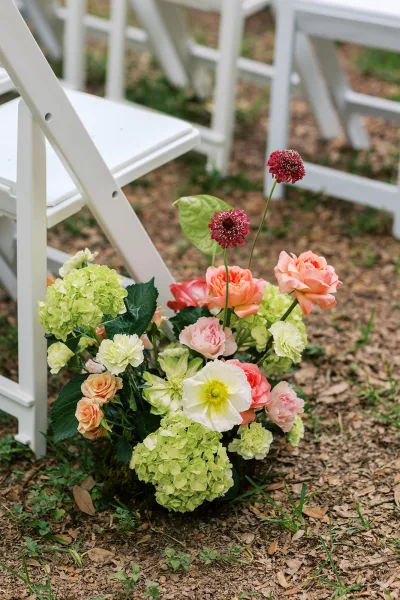 Ceremony aisle flowers serving as aisle marker flowers, nested in a moss base along a dirt ground scattered with fallen leaves and grass.