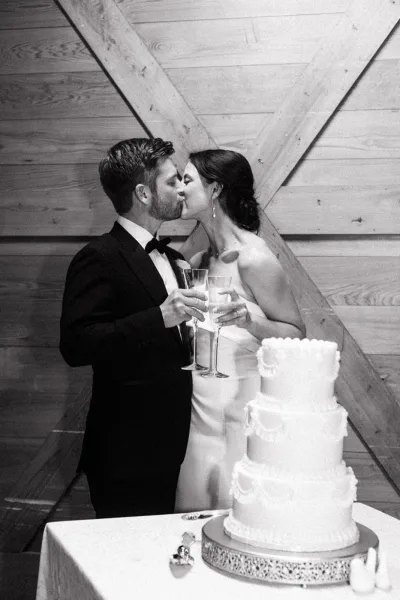 Wedding kiss portrait during a newlywed champagne toast, the couple clinks flutes against wood beams, capturing a warm indoor reception.