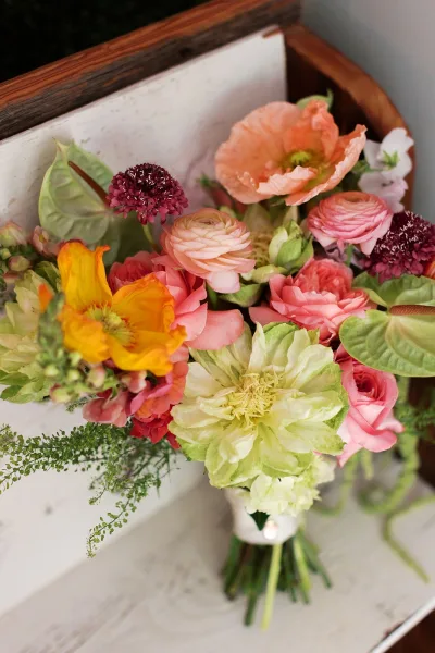 Bridal bouquet styled as a hand-tied bouquet, featuring poppies, resting on a wood tabletop against a white board backdrop.