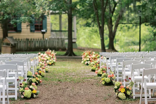 Ceremony aisle design with white folding chairs as garden ceremony decor on a grass lawn beneath garden trees.