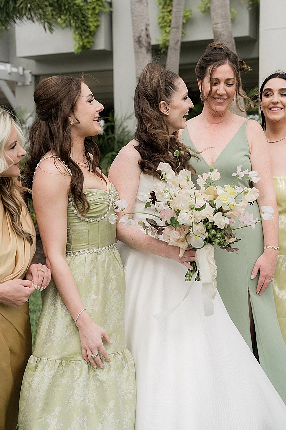 Bride with bridesmaids in sage green dresses laughing together, bride holding a bouquet with trailing ribbon outdoors by palm trees