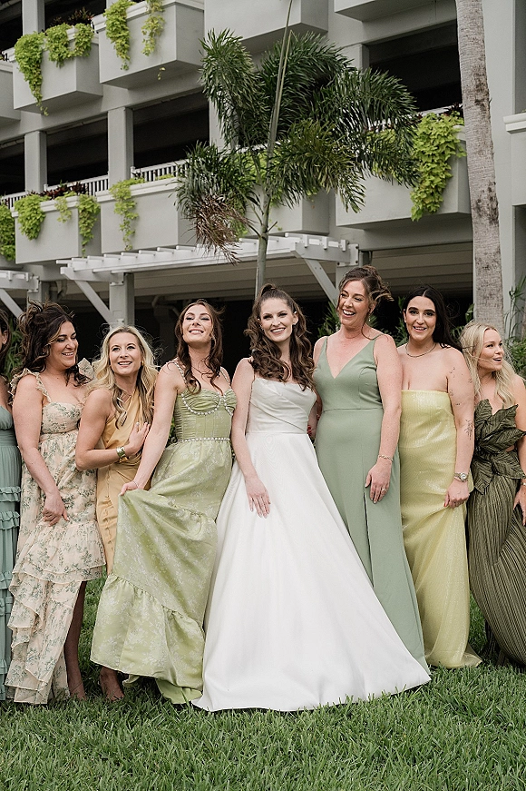 Bride with bridesmaids group photo, bride in strapless gown beside mismatched sage green dresses on a lawn with palm tree and hotel balconies