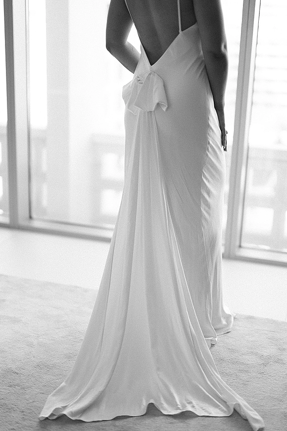 Wedding dress with a back bow and low back bridal gown silhouette, thin straps and train glowing in soft natural light by large windows