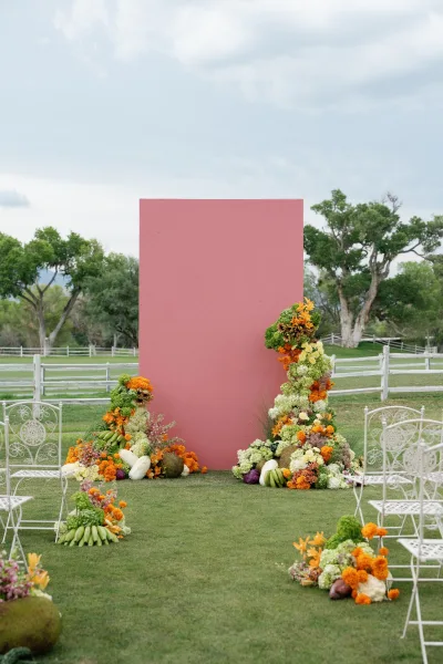 Ceremony backdrop featuring a pink panel altar edged with colorful florals, set on a lawn with a wooden fence and trees under a cloudy sky.