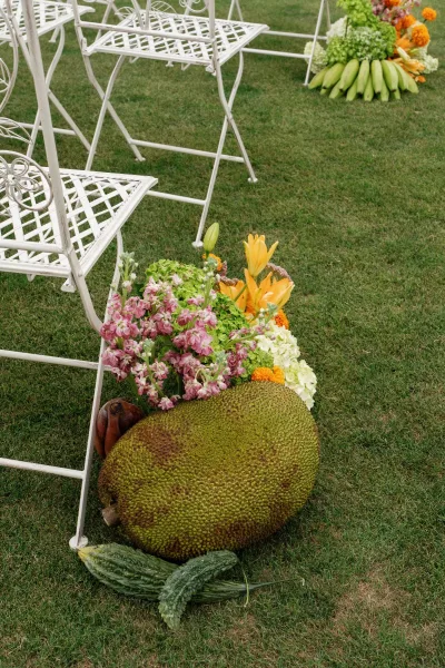 Ceremony aisle decor featuring an aisle marker arrangement with jackfruit nestled among vibrant petals beside each chair.