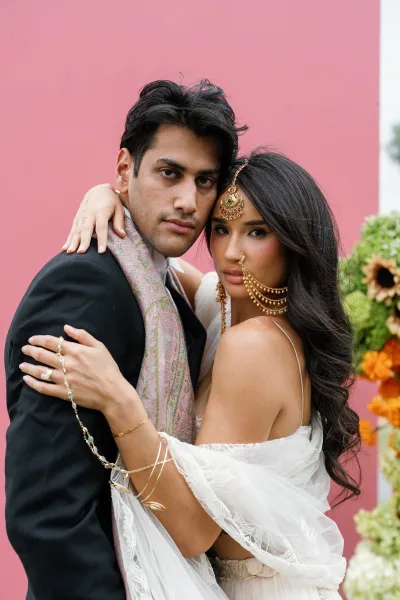 Couple portrait of a bride and groom in a close embrace, the bride wearing a maang tikka headpiece against a pink floral backdrop.