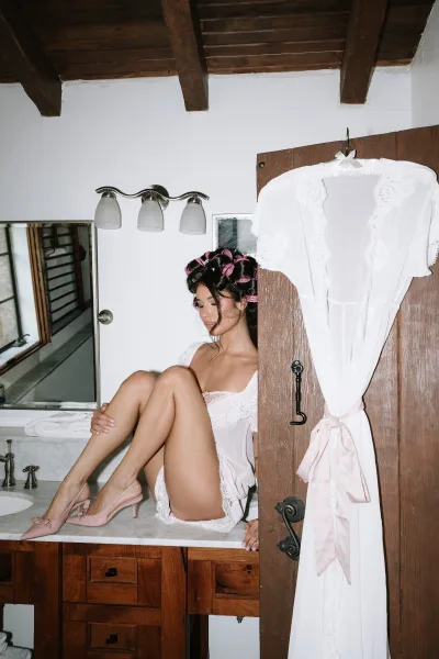 Bridal getting ready scene shows a bride in rollers adjusting a lace-trim robe at a bathroom vanity mirror with warm sconce lights.