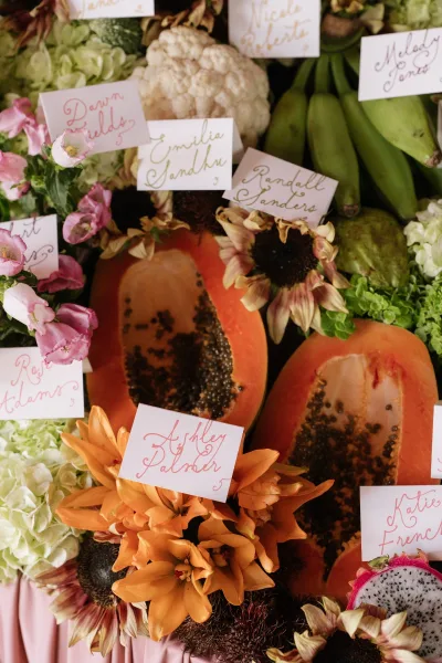 Escort card display featuring fruit place cards set in papaya, arranged neatly on pink table linen atop a tabletop.