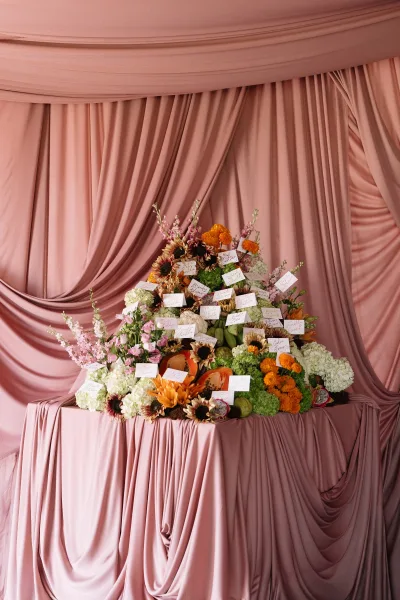 Escort card display featuring floral escort cards arranged with papaya accents before a pink drapery backdrop.