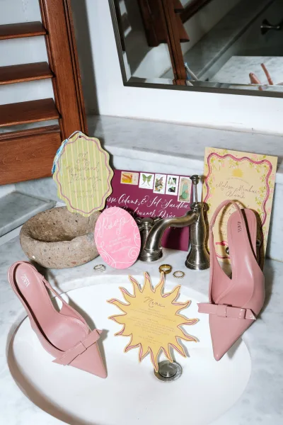 Bridal shoes—blush wedding shoes—set on a marble bathroom counter beside wedding rings, beneath a mirror.