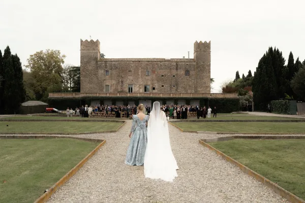 Wedding processional with bride walking to ceremony in a long-sleeve gown and veil, carrying a bouquet along a gravel aisle at a stone castle courtyard