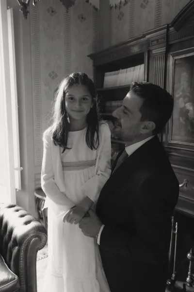 Father daughter portrait of a man in suit and tie holding a girl in a white dress and cardigan, posed by a bookshelf in window light