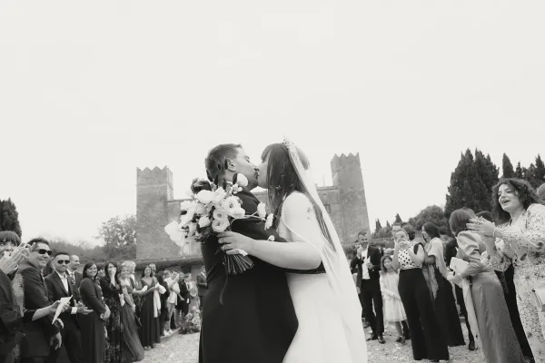 Wedding kiss as the bride and groom embrace, veil trailing and bouquet in hand, while guests cheer in a castle courtyard outdoor ceremony
