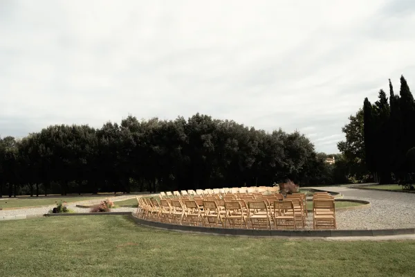 Ceremony seating with wood folding chairs in a curved layout, with floral arrangements and pampas grass accents on a lawn by gravel paths