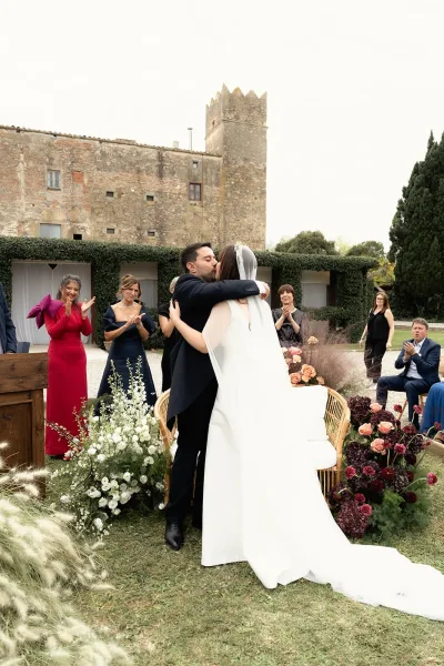 Ceremony kiss moment as bride and groom embrace, her long veil and train flowing beside floral arrangements in a stone courtyard setting