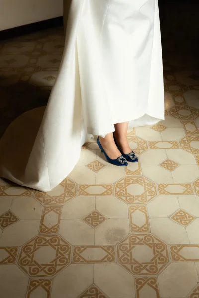 Bridal shoes, blue wedding heels with jeweled buckles peeking beneath the gown hem on a patterned tile floor in soft shadowed light