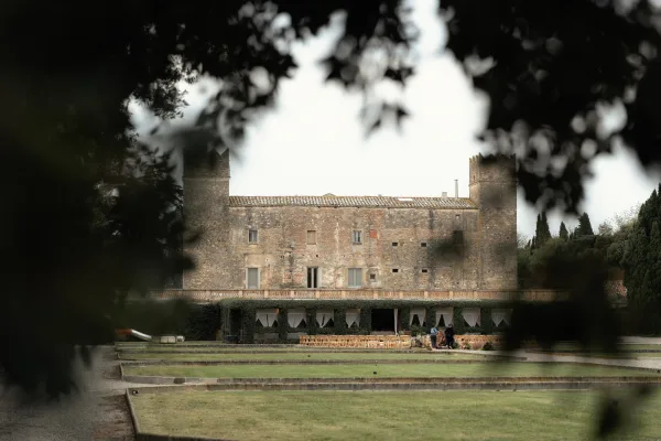 Outdoor reception setup with long banquet tables and wooden chairs, greenery garlands and white draping in a stone manor courtyard lawn
