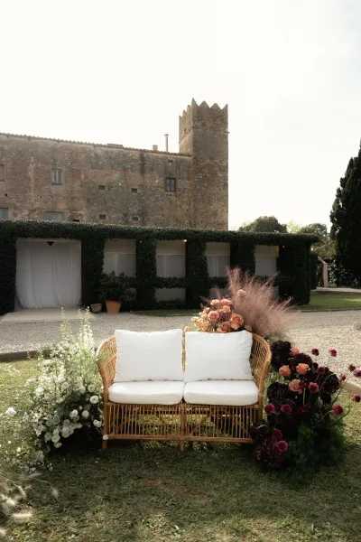 Wedding lounge seating with a rattan loveseat and white cushions, framed by rose florals and pampas grass on a lawn by a stone tower