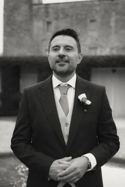 Groom portrait in a tailored three-piece suit with a white rose boutonniere, posed in a stone courtyard with hedges behind