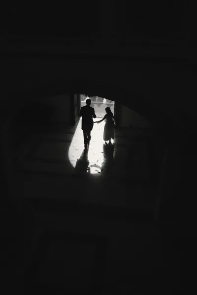 Wedding exit moment as newlyweds walking away hand in hand, bride in gown and groom in suit, silhouetted in a sunlit doorway