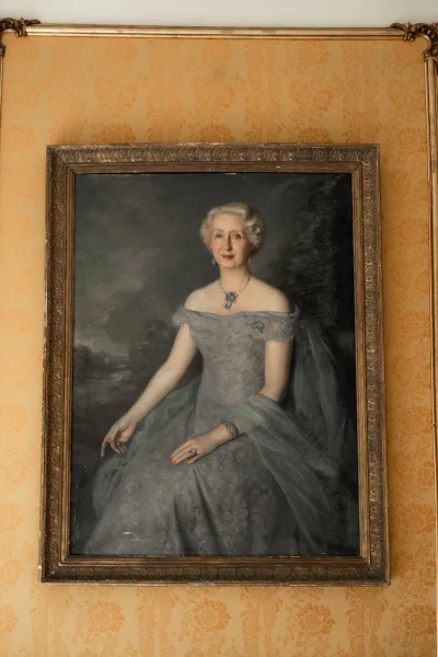 Framed portrait painting in an ornate gold frame showing a woman in an evening gown with statement necklace against damask wallpaper and molding