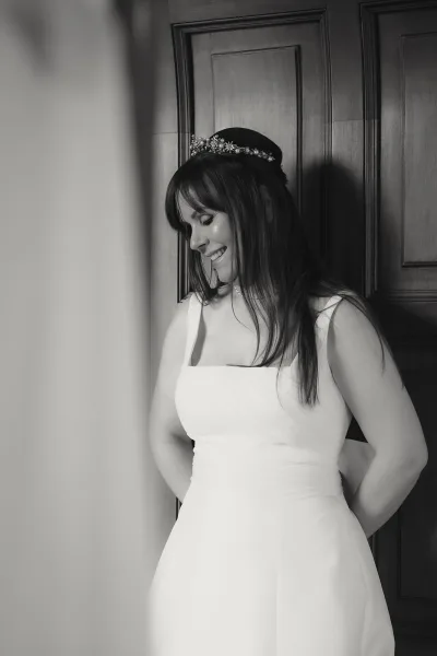 Bridal portrait in black and white of a smiling bride looking down, wearing a tiara and necklace beside a wood paneled door indoors