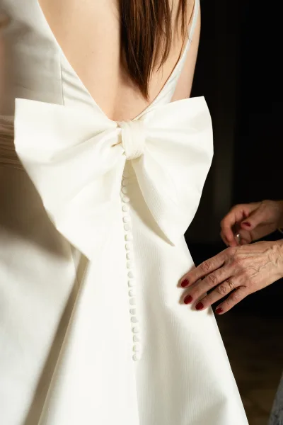 Wedding dress detail with oversized bow as hands fasten fabric-covered buttons, engagement ring and red nails against a dark indoor backdrop