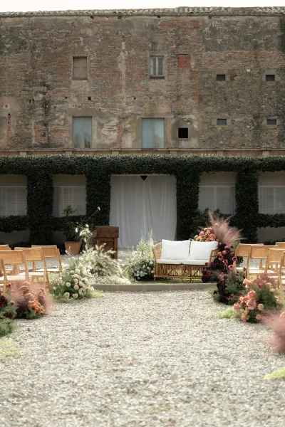 Ceremony setup with outdoor ceremony aisle, wood chairs flanking a gravel path lined with florals and pampas before white drape in brick courtyard