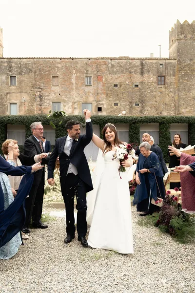 Wedding recessional as bride and groom raise hands, bouquet and veil flowing while guests toss petals in an ivy-lined stone courtyard