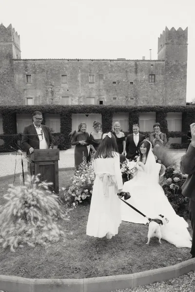 Ceremony moment at an outdoor wedding ceremony with bride in veil holding bouquet as guests laugh in a stone courtyard setting