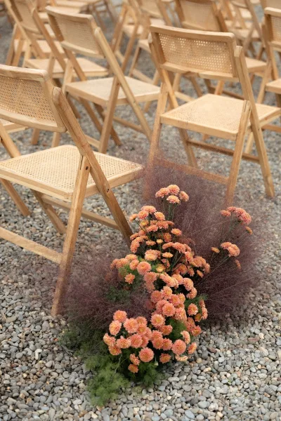 Ceremony aisle decor with an aisle flower arrangement of peach blooms, ornamental grass, and greenery beside wood folding chairs on gravel