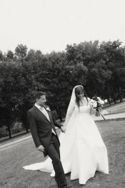Couple portrait of bride and groom walking hand in hand, bride in strapless dress and veil with bouquet, along tree-lined path